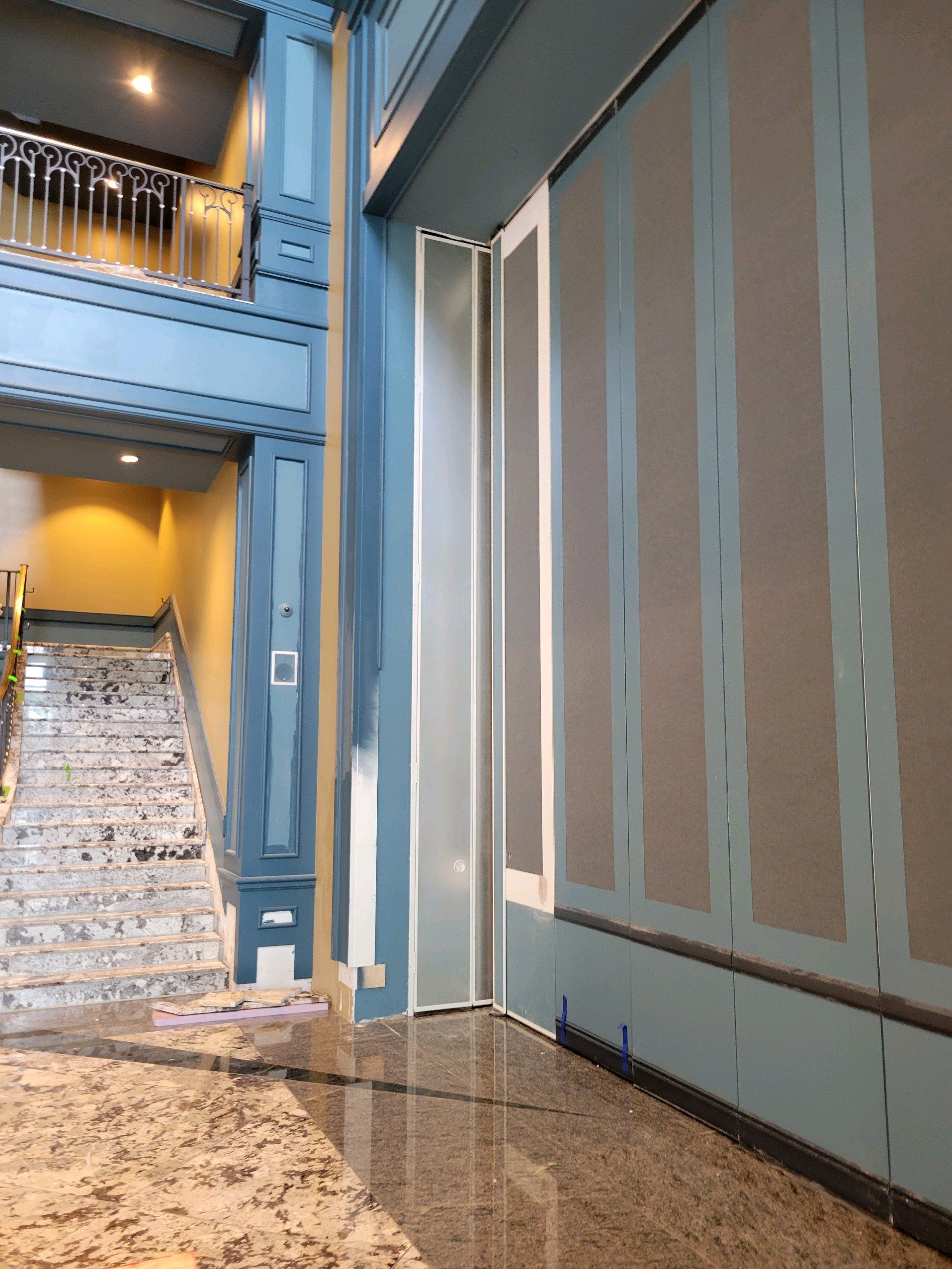 An interior shot of a lobby. Blue painted trim surrounds the hallway and staircase with a marbled floor.