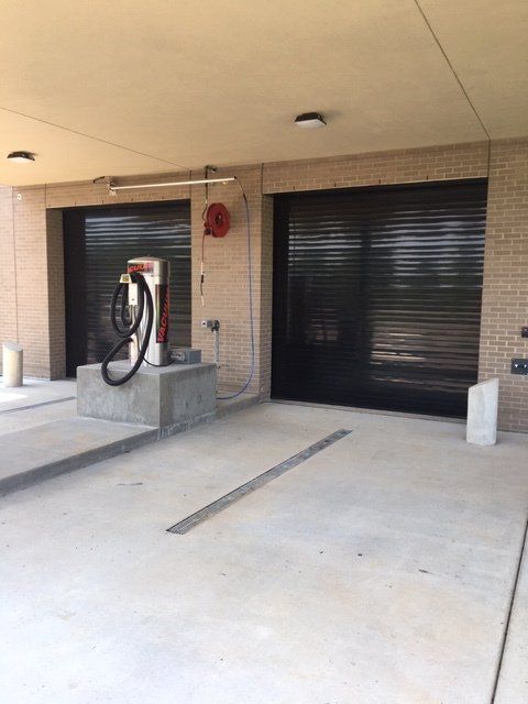 Electric vehicle charging station with black roller doors, concrete platform, and hose reel.