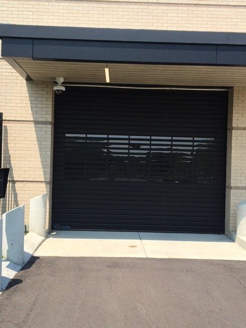 Black roll-up garage door with a clear window section, under a beige brick building with security camera.