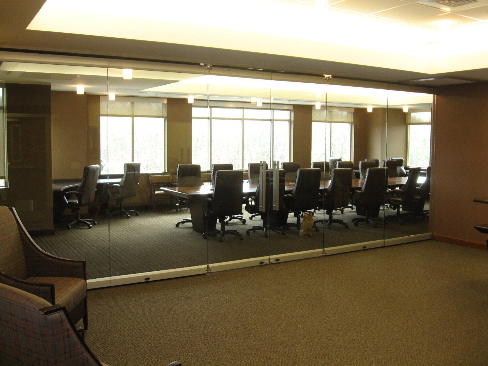 Conference room with large table and chairs behind a glass wall. Windows in the background.