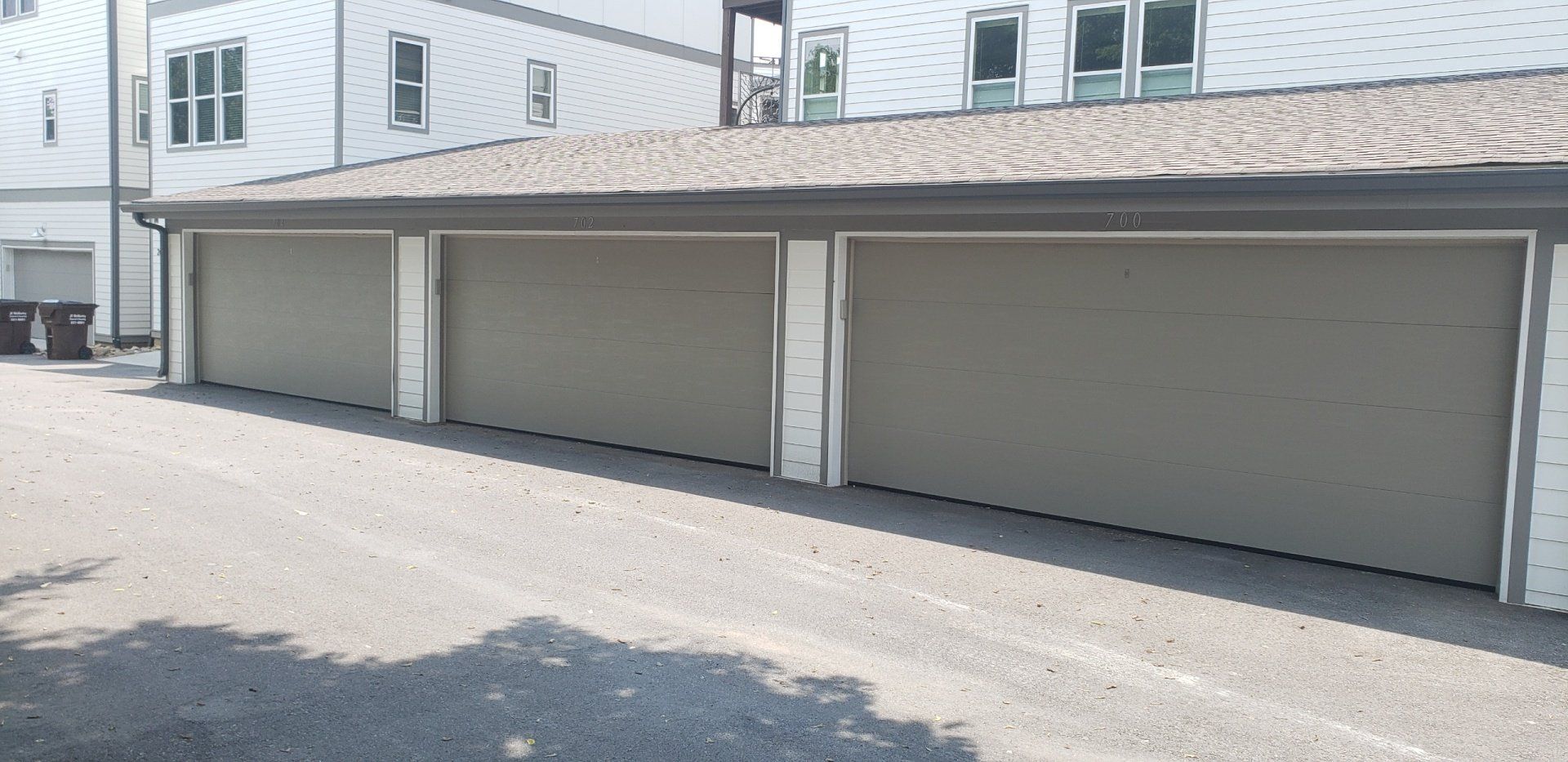 Three attached garage doors, tan color, on a gray gravel driveway, with a white building in the background.