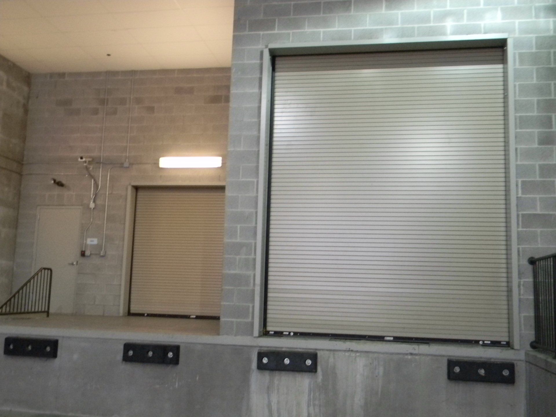 Loading dock with two large, closed, rolling doors made of metal. Gray brick walls.