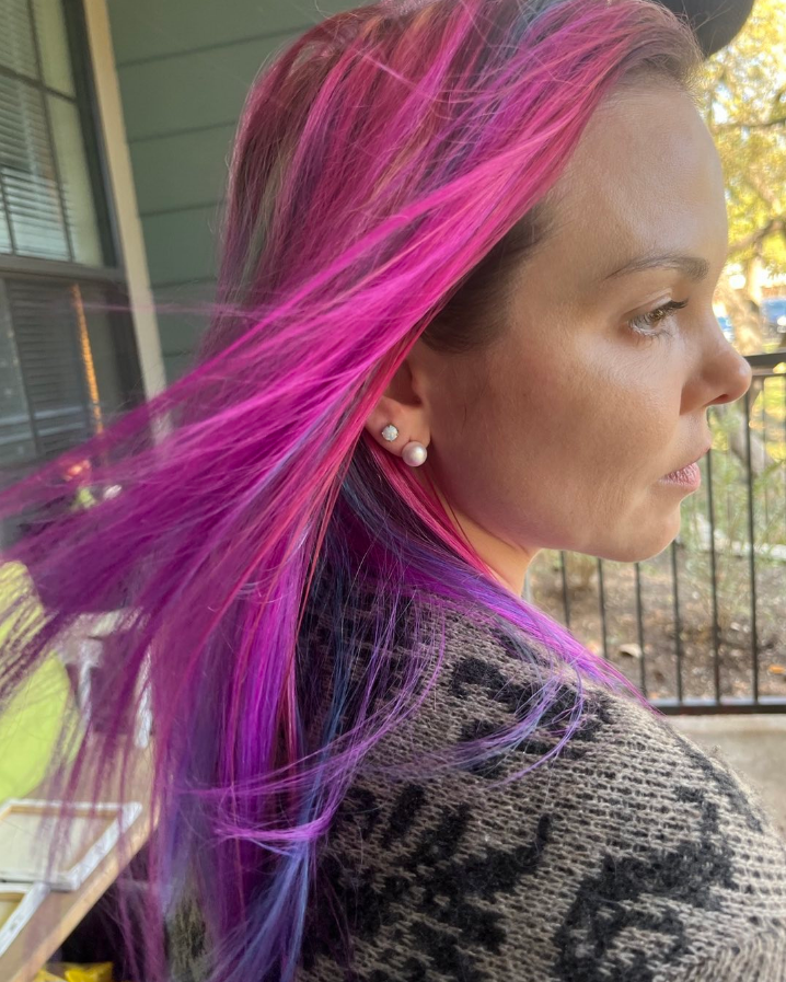 Woman with long, pink and purple hair; wearing earrings and a patterned sweater. Outside.