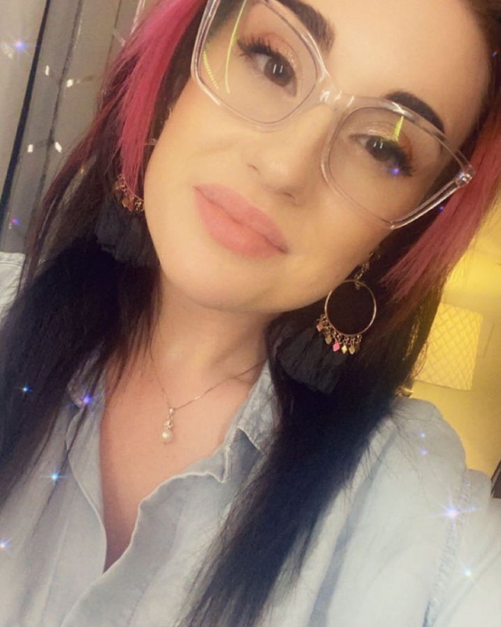Woman with pink-streaked dark hair, clear glasses, and light makeup smiles. Wearing earrings, necklace.