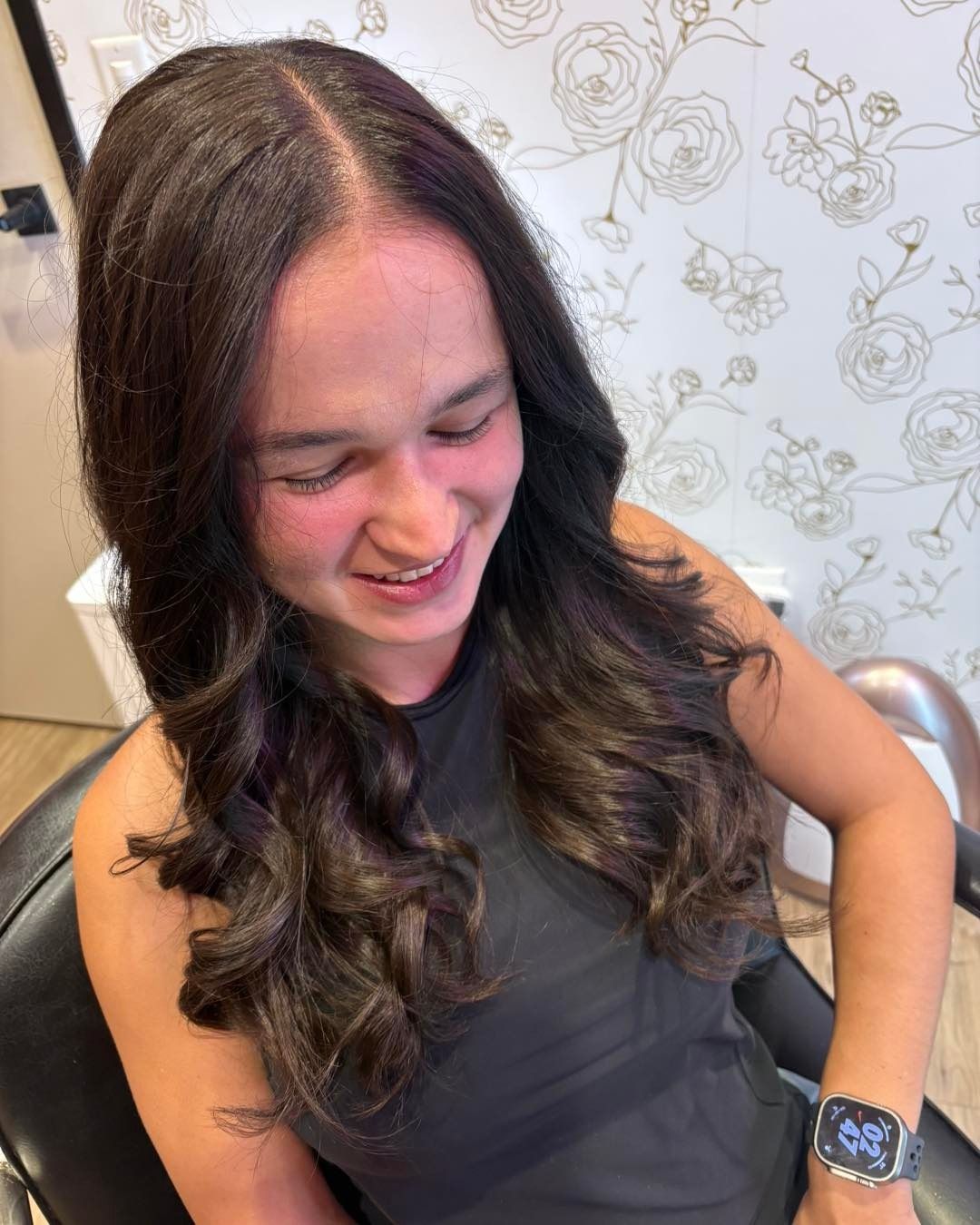 Woman with long, wavy brown hair, smiling, in a salon chair. Wearing a black top.