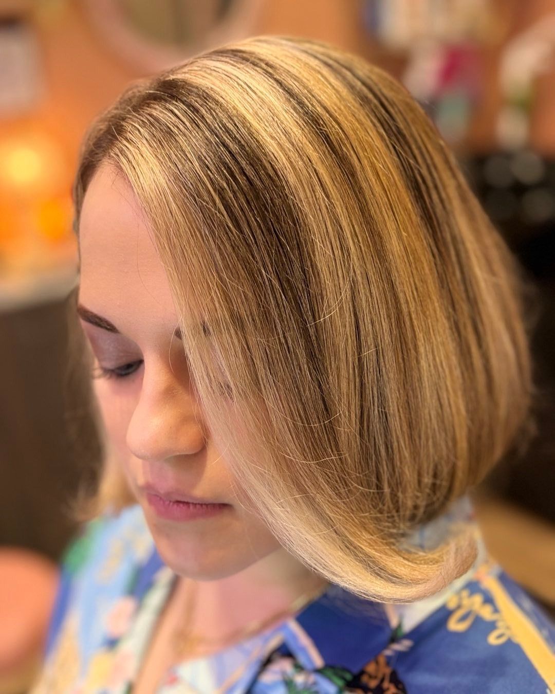 Woman with short, layered blonde hair, highlights, and a blue floral top, looking down.