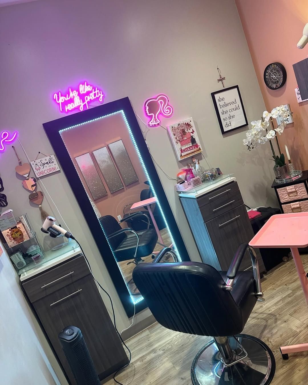 Hair salon interior with mirror, chair, neon signs, and decorative items in pink and gray tones.