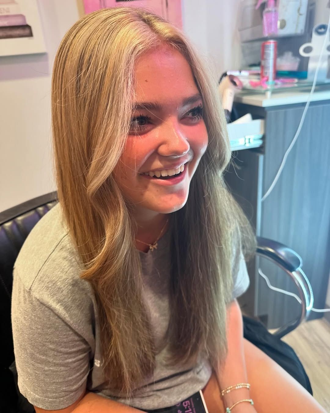 Blonde-haired person smiles, sitting in a salon chair. They are wearing a gray shirt with a light-filled background.