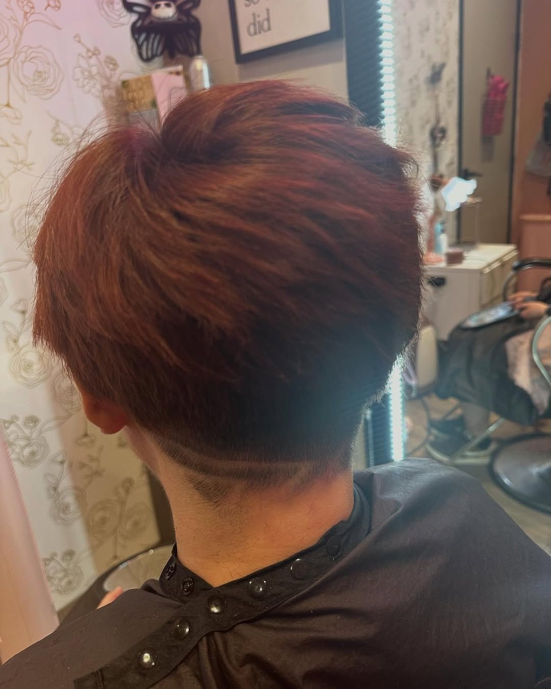 Person with short, reddish-brown hair in a salon. The back of the neck has a shaved design.