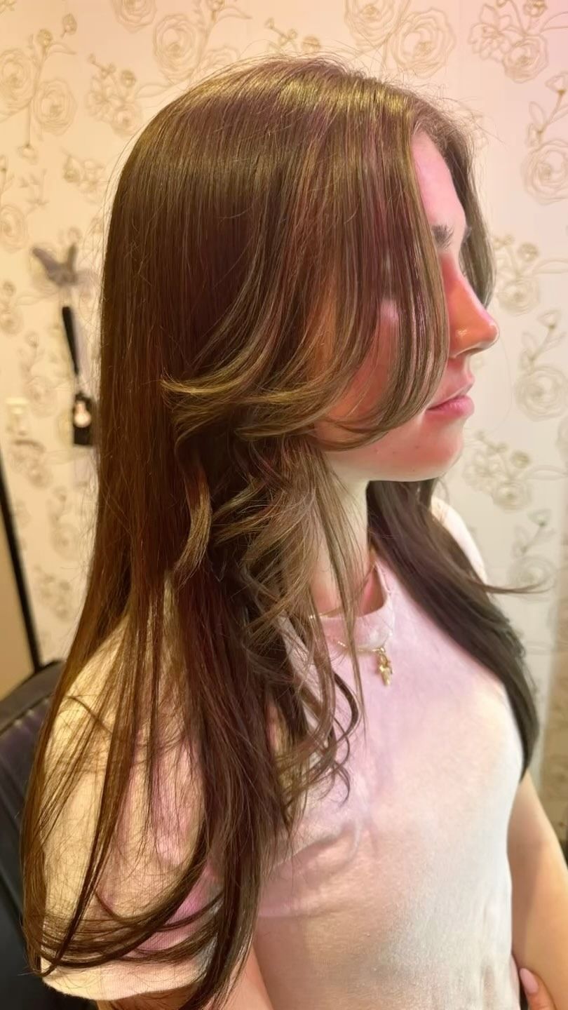 Woman with long, layered brown hair, some lighter sections, in a salon setting, profile view.