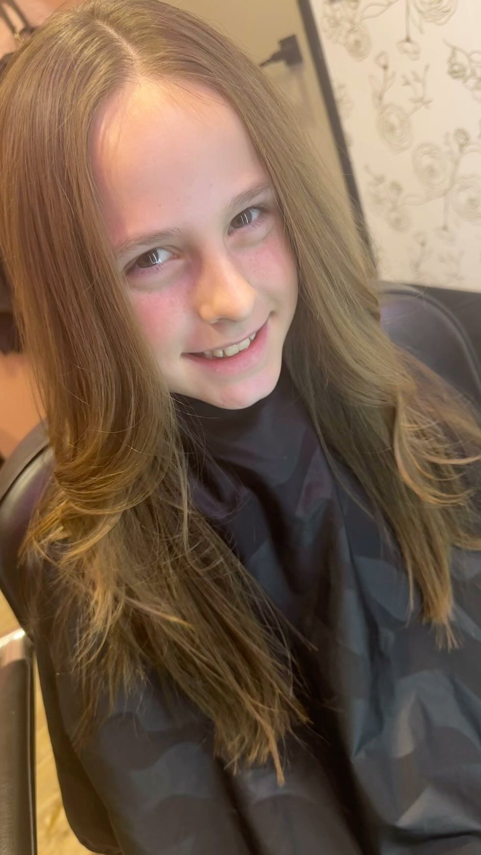 Young person with long, layered hair smiling, wearing a salon cape.