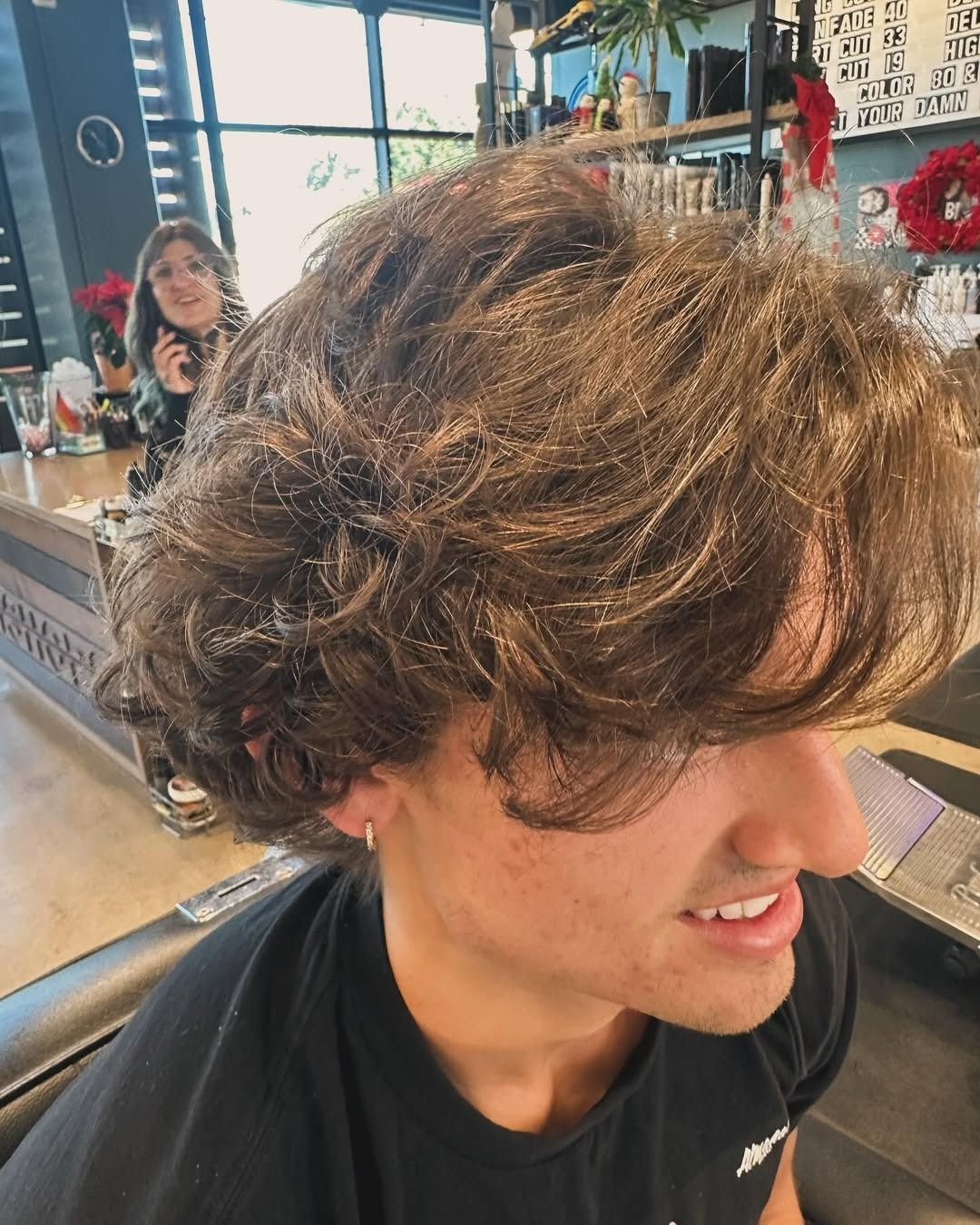 A person with wavy brown hair, smiling, in a salon. A person is visible in the background.