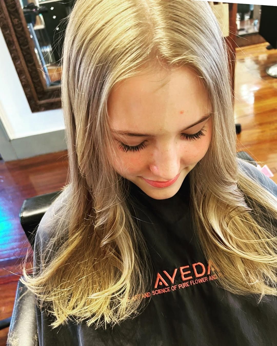 Girl with long blonde hair, smiling in a salon. Hair is layered and styled, wearing an Aveda smock.