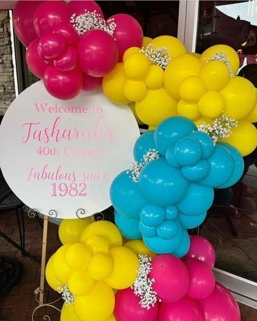 A sign is surrounded by colorful balloons and flowers.