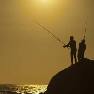 Two men are fishing on a rock near the ocean at sunset.