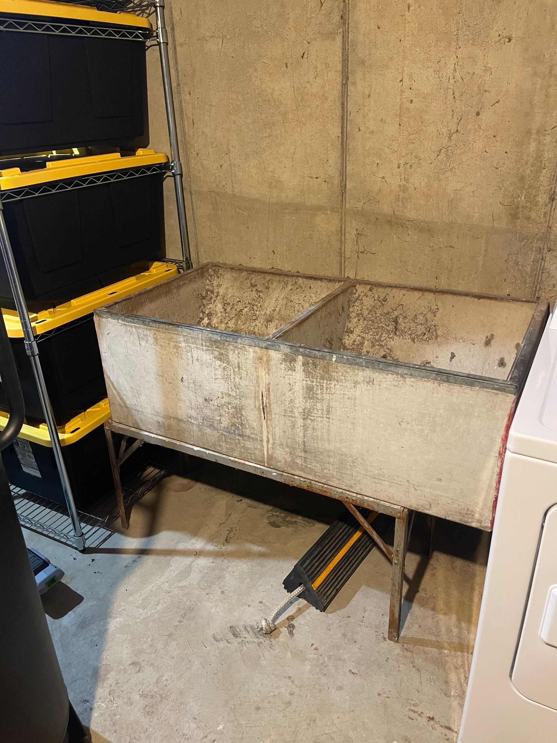 A worn, double basin laundry sink in a basement. Beside it, a black shelving unit with yellow bins.