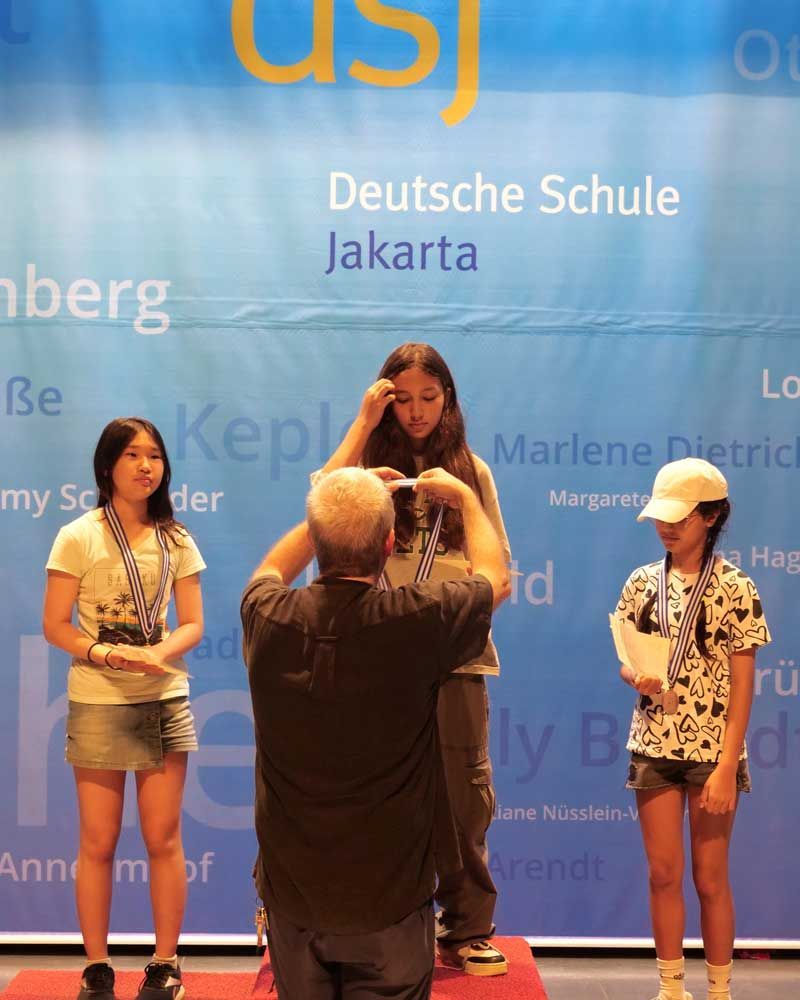 A man is taking a picture of three girls in front of a sign that says deutsche schule jakarta