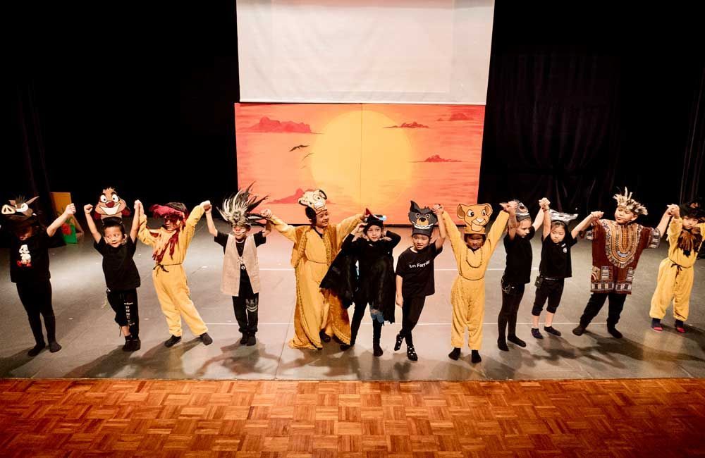 A group of children in costume are standing on a stage holding hands.