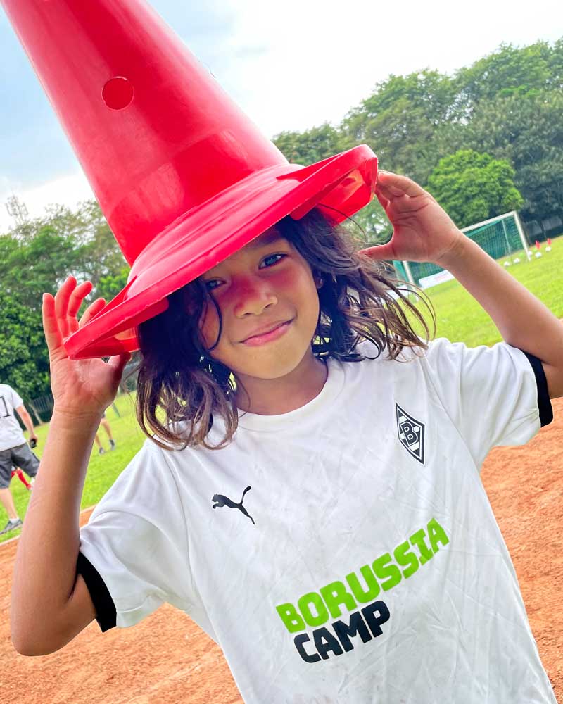 Smiling child wearing a Borussia Camp shirt, playfully placing a red traffic cone on their head