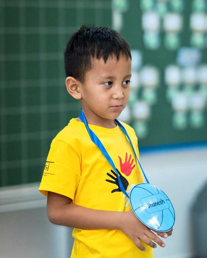 A young boy wearing a yellow shirt with a hand on it is standing in front of a blackboard.