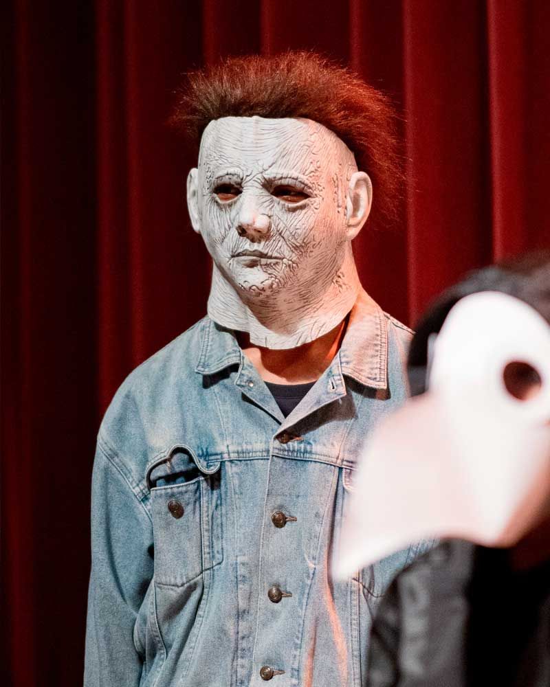A man wearing a mask and a denim jacket is standing in front of a red curtain.
