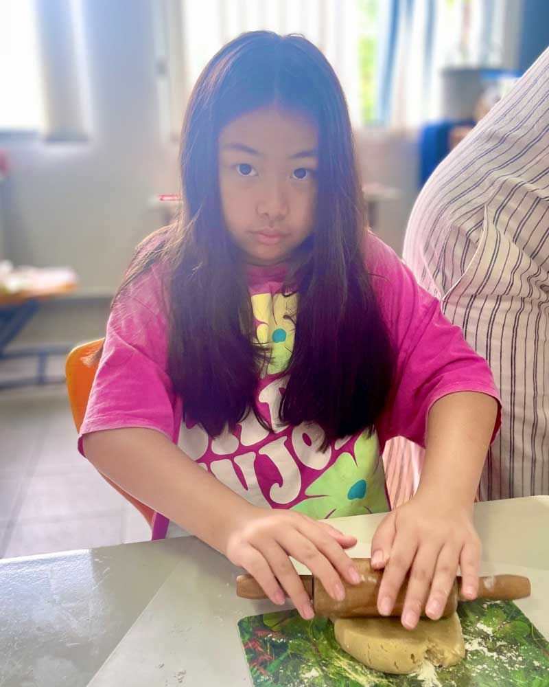 A young girl is sitting at a table rolling dough with a rolling pin.