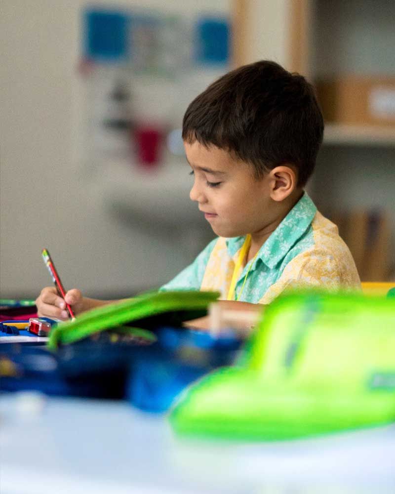 A young boy is sitting at a table in a classroom writing in a notebook.