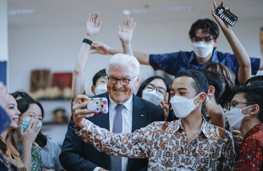 A man in a suit and tie is taking a selfie with a group of people wearing masks.