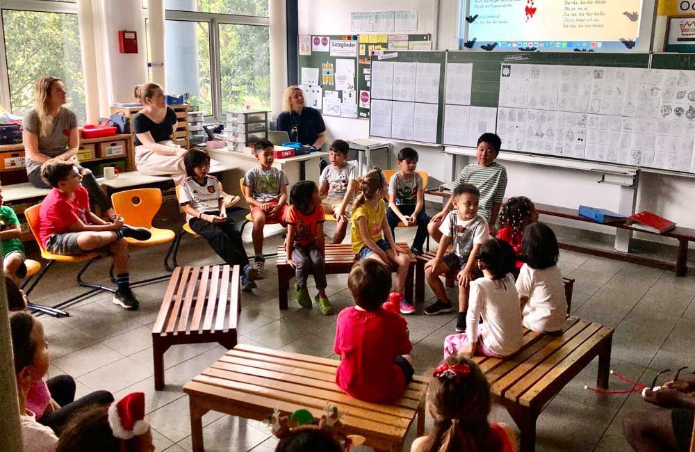 A group of children are sitting around wooden benches in a classroom.