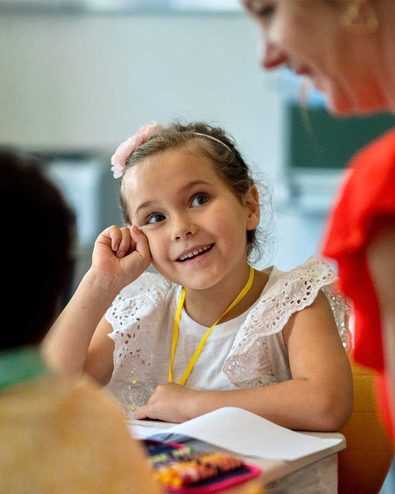 A little girl is sitting at a desk in a classroom with a teacher.