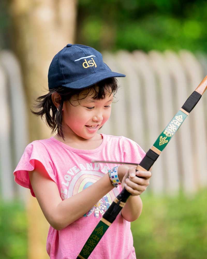 Smiling girl in a pink shirt and cap holding a toy bow and arrow, ready to aim.
