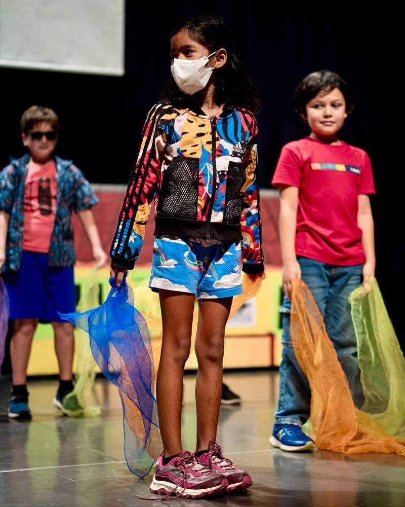 A girl wearing a mask is standing on a stage with other children.