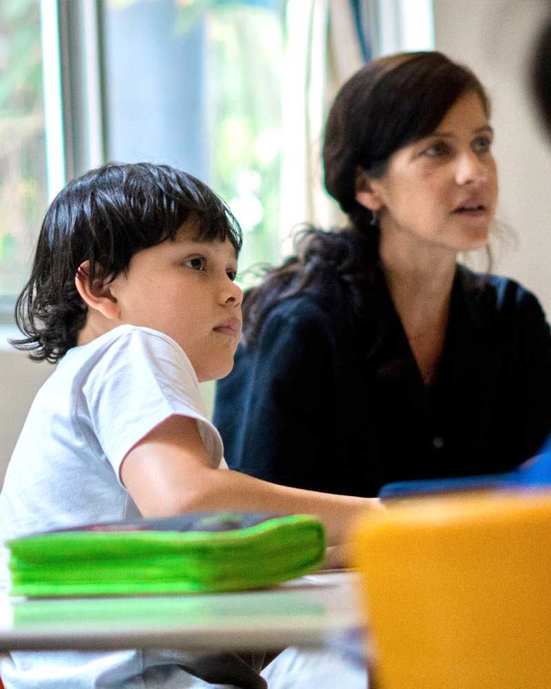 A woman and a child are sitting at a desk in a classroom.