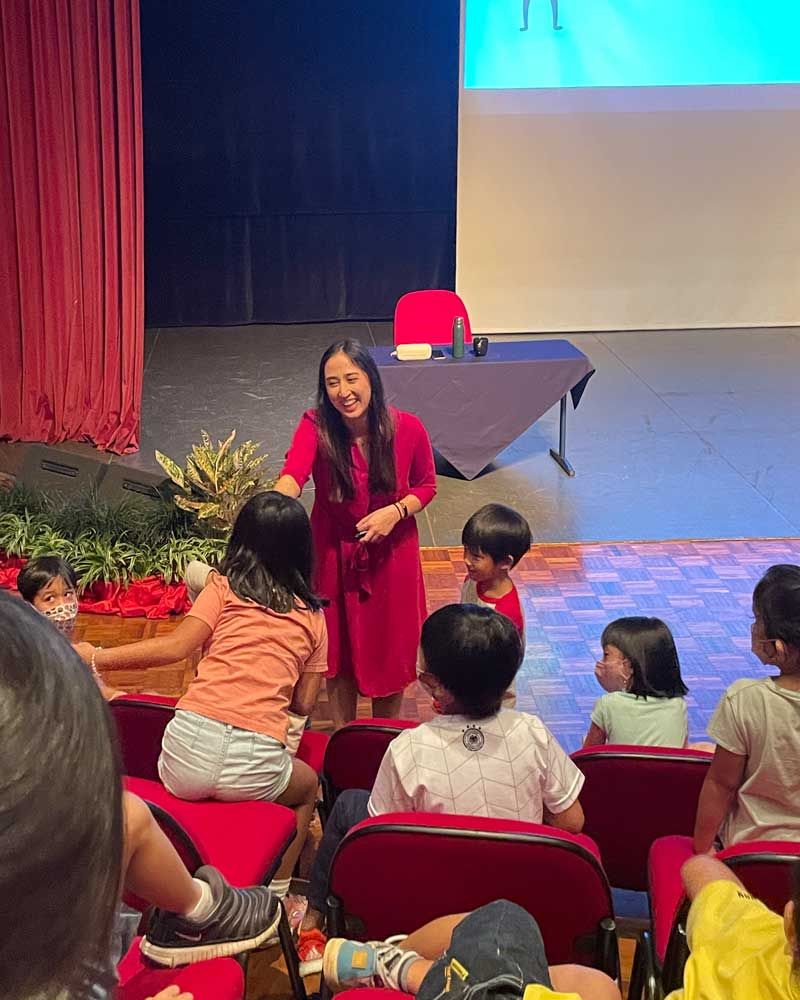 A woman is standing in front of a group of children in an auditorium.