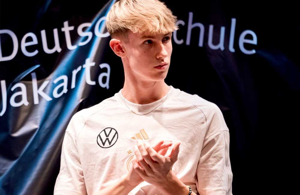A young man is standing in front of a sign that says deutsch schule jakarta.