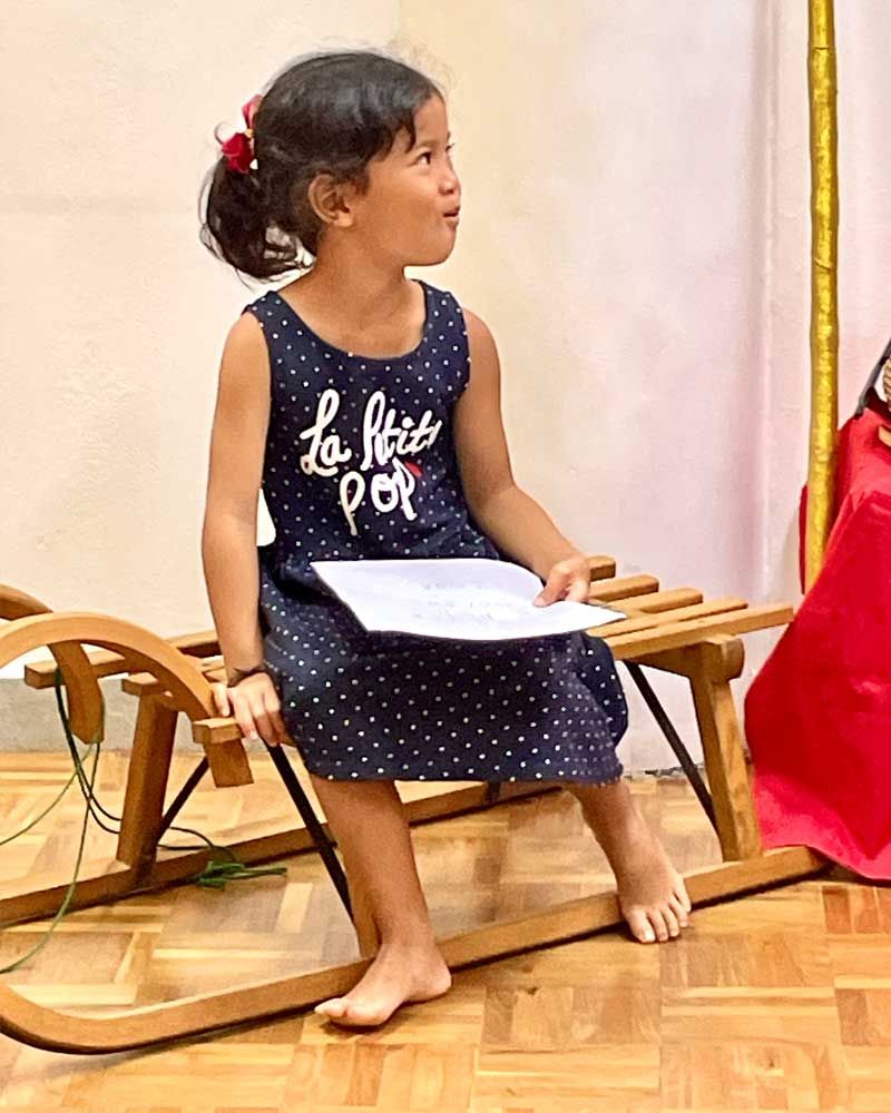 A little girl in a blue dress is sitting on a wooden sled holding a piece of paper.