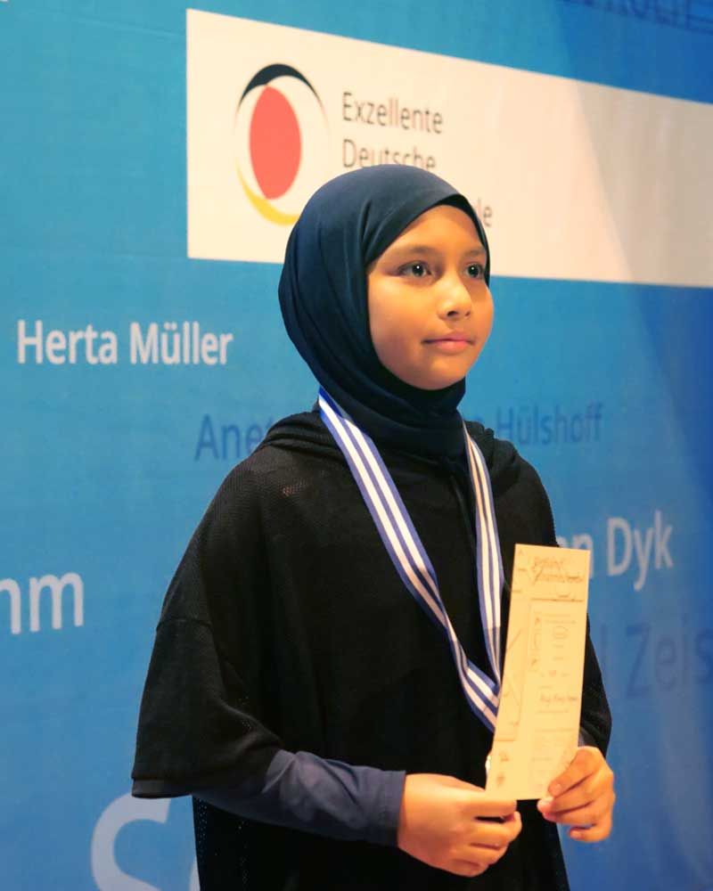 A young girl wearing a hijab and a medal stands in front of a sign that says herta muller