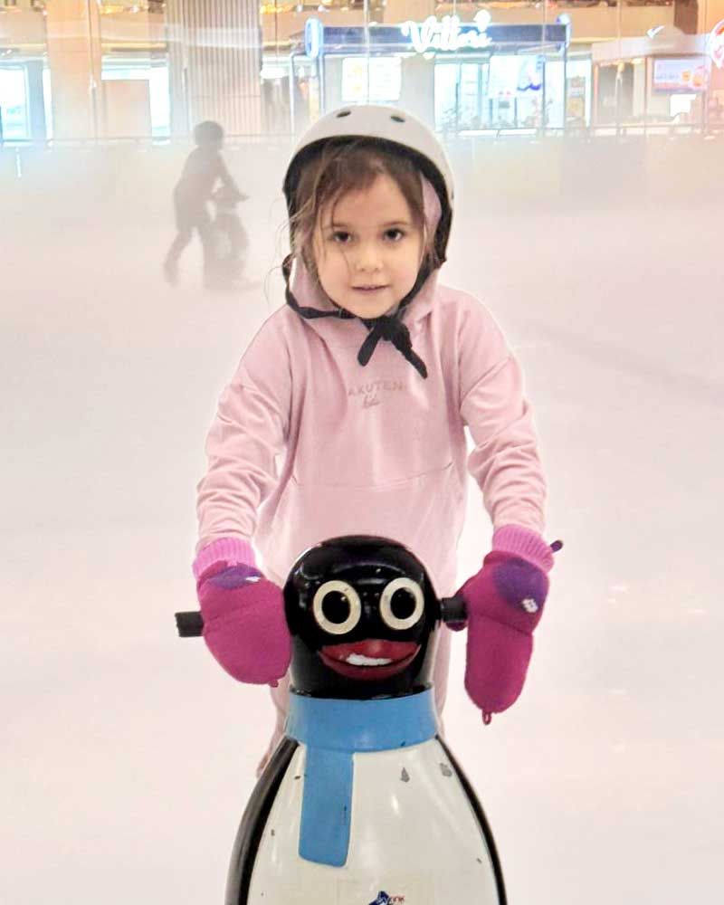 A little girl is riding a penguin scooter on an ice rink.