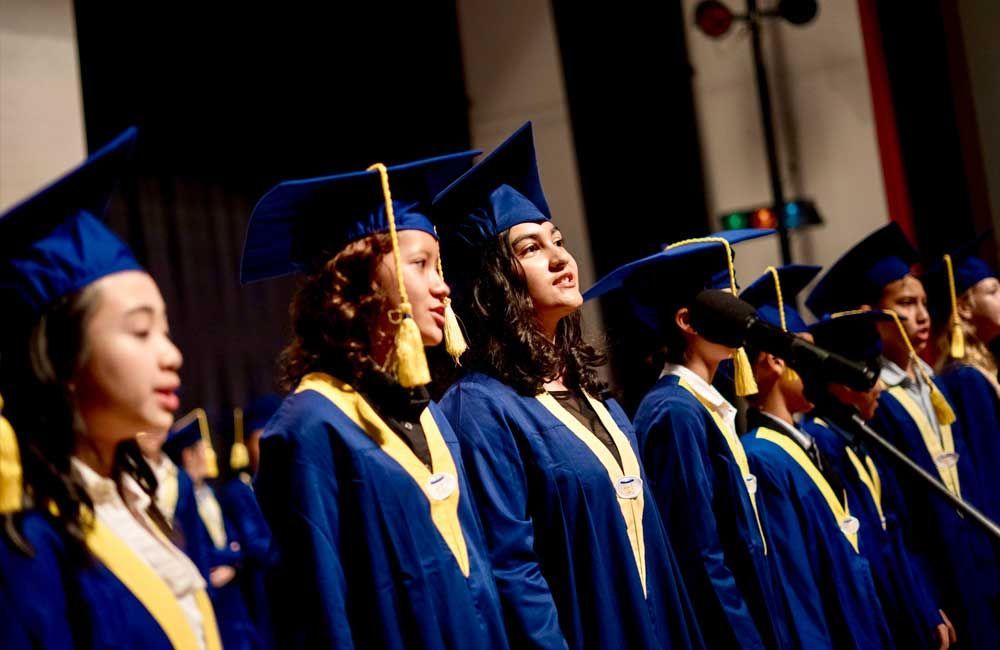 A group of graduates are singing into microphones on a stage.