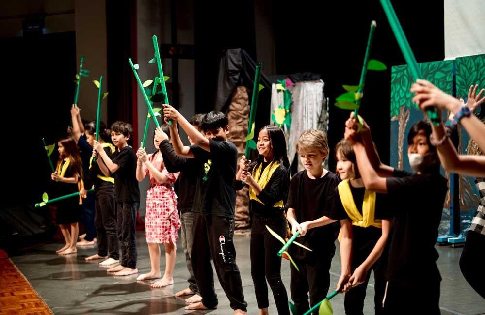 A group of children are standing on a stage holding green sticks.