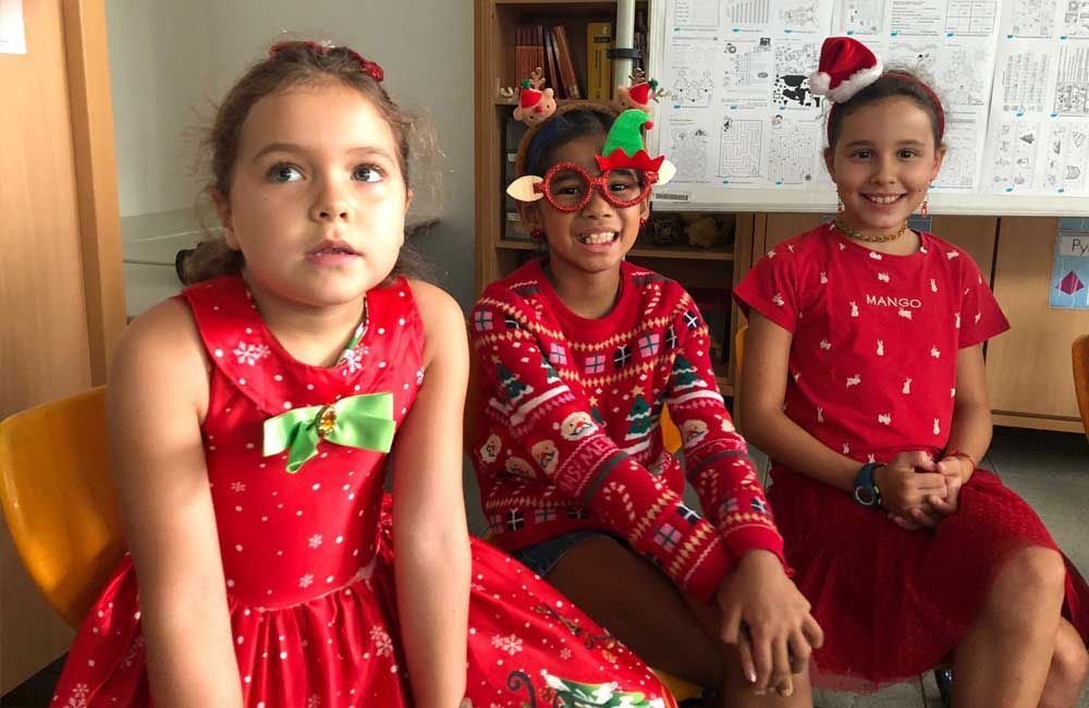 Three little girls are sitting next to each other wearing christmas sweaters and dresses.