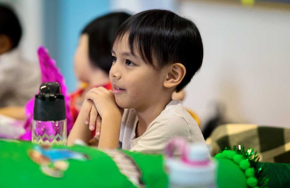 A young boy is sitting at a table with his hands on his chin.