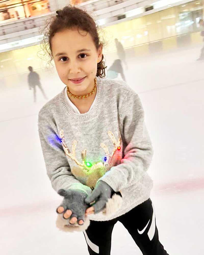 A little girl wearing a sweater and gloves is standing on an ice rink.