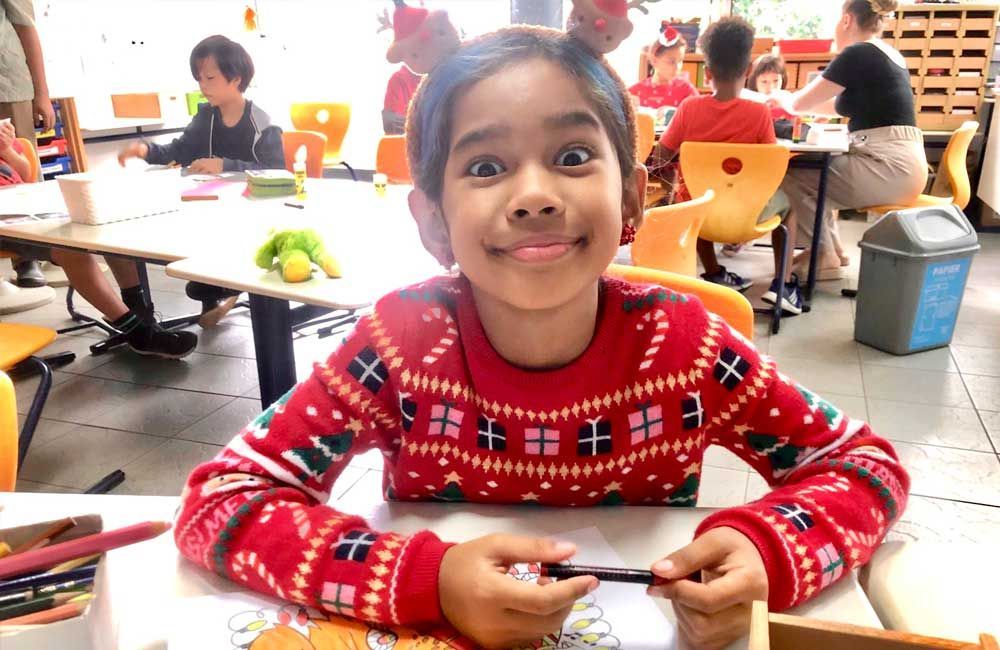 A young girl wearing a christmas sweater and reindeer antlers is sitting at a table.