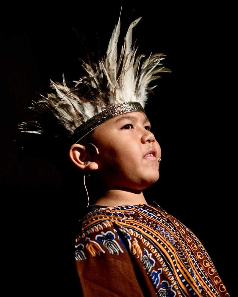 A young boy is wearing a feathered headdress and a brown shirt.