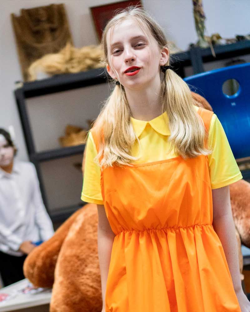A girl in an orange dress and yellow shirt is standing next to a teddy bear.