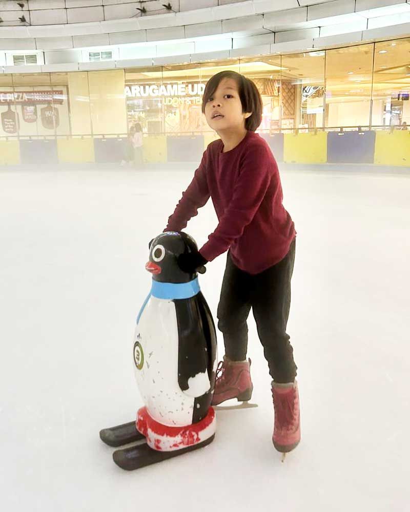 A young boy is standing next to a penguin statue on an ice rink.
