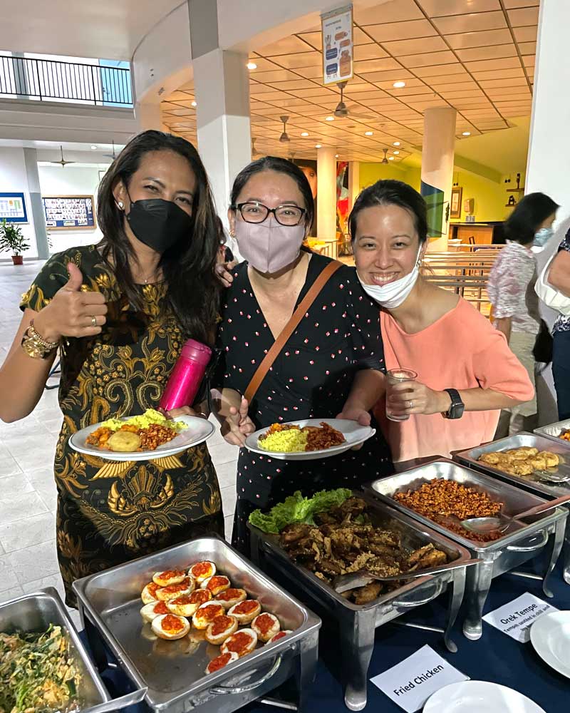 Three women wearing face masks are standing in front of a buffet table holding plates of food.