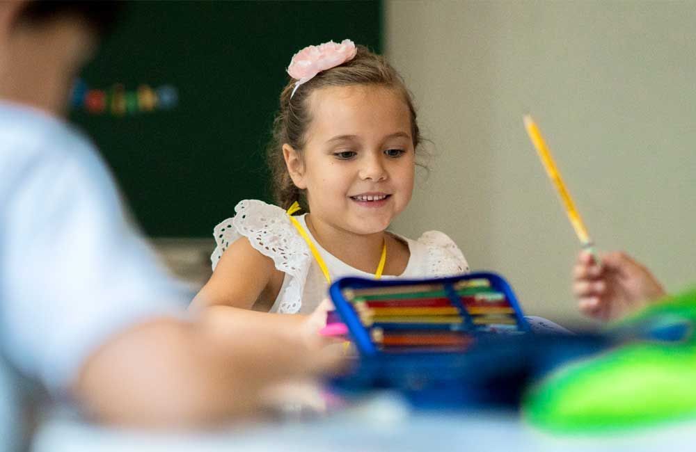 A little girl is sitting at a desk in a classroom holding a pencil.