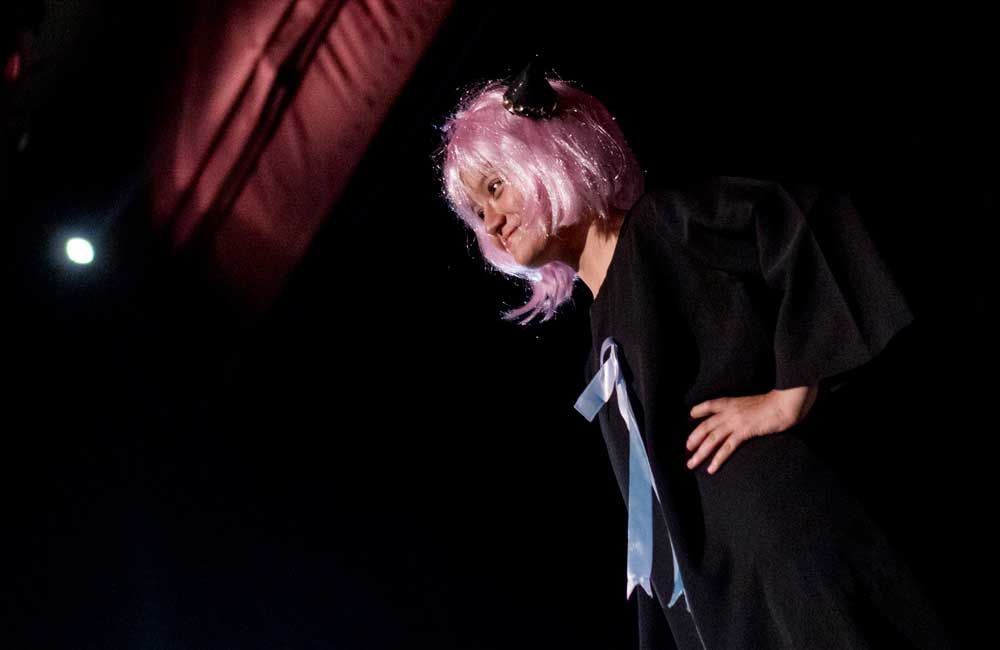 A woman wearing a pink wig and a black cape is standing on a stage.